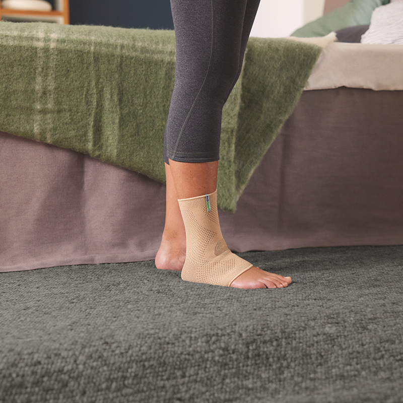 Woman wearing leggings and an ankle support stands on a plush carpet in a bedroom 